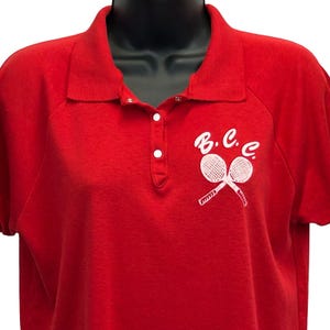 Puede incluir: Un polo rojo con mangas cortas y cuello. La camisa presenta un gráfico blanco de raquetas de tenis cruzadas y las letras "B.C.C." en el pecho. La camisa tiene una tapeta con botones y un corte clásico.