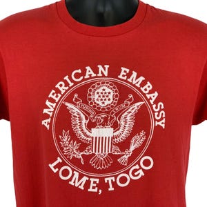 Op de afbeelding: Rood t-shirt met een witte afbeelding van de Amerikaanse ambassade in Lomé, Togo. Het ontwerp bevat een adelaar, sterren en de tekst "AMERICAN EMBASSY" en "LOME, TOGO" in een cirkelvormige opstelling. Het shirt is gemaakt van zacht materiaal.
