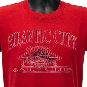 May include: A red short-sleeved t-shirt featuring a distressed graphic print. The text reads ATLANTIC CITY SAIL CLUB. The design includes a diamond-shaped frame containing a sailboat illustration, flanked by two small ship wheel icons.
