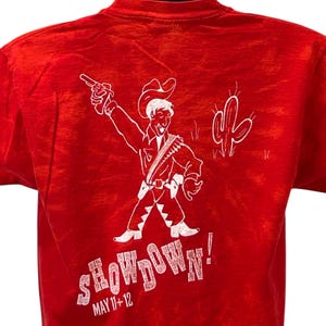 Puede incluir: Camiseta roja con un dibujo blanco de un vaquero con una pistola, un cactus y la palabra "SHOWDOWN!" con la fecha "MAY 11+12". La camiseta tiene un aspecto vintage y desgastado.