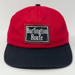 May include: A red baseball cap with a black brim and a black button on top. The cap features a rectangular patch with the words "Burlington Route" in white lettering on a black background.