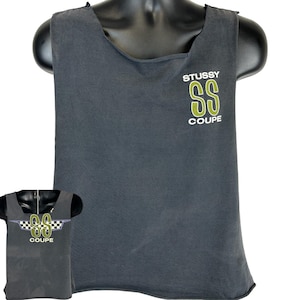 Puede incluir: Una camiseta sin mangas gris oscuro con el texto "STUSSY" encima de un logotipo "SS" verde y la palabra "COUPE" debajo. La camiseta se muestra en un maniquí negro.