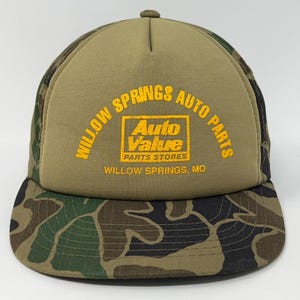 Może przedstawiać: Zielono-brązowa czapka typu trucker w kamuflażu z żółtym napisem "Willow Springs Auto Parts". Czapka posiada również logo "Auto Value Parts Stores" i miasto "Willow Springs, MO".