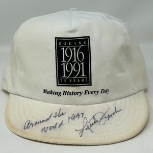 Könnte beinhalten: Weißes Baseball-Cap mit dem Boeing-Logo und den Jahren 1916-1991, zum 75-jährigen Jubiläum. Der Text "Making History Every Day" steht darunter. Auf dem Schirm befinden sich handschriftliche Texte und eine Unterschrift.