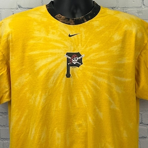 May include: Yellow tie-dye t-shirt with a black collar and a small Nike logo. The shirt features a Pittsburgh Pirates logo on the front. The logo is a black letter "P" with a skull and crossbones design.