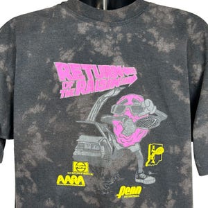 May include: Dark gray tie-dye t-shirt with a graphic featuring a pink raisin character playing racquetball. The text "RETURNS OF THE RAISINS" is above the character. Logos for Pepsi, AARA, and Penn Racquetball are also visible.