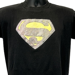 Puede incluir: Camiseta negra con el logo de Superman. El logo tiene forma de diamante con una 'S' amarilla dentro de un contorno plateado. La camiseta tiene cuello redondo y mangas cortas, y está hecha de un material suave.