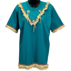 Peut inclure: T-shirt de couleur turquoise avec un col rond et des manches courtes. La chemise est ornée d'une bordure de franges crème le long de l'encolure, des bords des manches et de l'ourlet. La frange est accentuée d'une fine rayure rouge. Des ornements argentés décorent l'encolure.