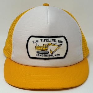 May include: A yellow and white trucker cap with a curved peak. The front panel is white with a black and yellow patch that reads "K.W. PIPELINE, INC. HENDERSON, NEV." The cap has a yellow mesh back and a yellow button on top.