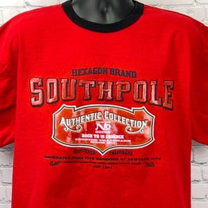 May include: A red t-shirt with a black collar featuring the text "SOUTHPOLE" in large, outlined letters. Below, a graphic reads "AUTHENTIC COLLECTION" and "BACK TO ESSENCE". The shirt also includes the text "HEXAGON BRAND".