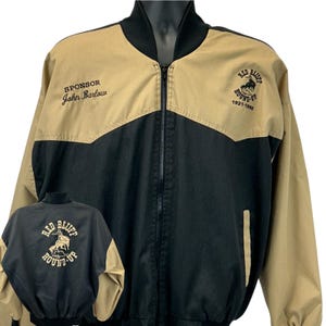 Op de afbeelding: Een zwart en bruin jack met een zwarte kraag en rits. De jas heeft de tekst "SPONSOR John Barlow" op de voorkant en "RED BLUFF ROUND-UP 1921-1986" op de voor- en achterkant. Een jas in vintage stijl.