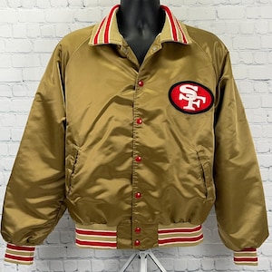May include: A gold satin bomber jacket featuring a red and white striped collar and cuffs. The jacket has a front snap button closure and a prominent oval patch on the left chest displaying the red and white San Francisco 49ers team logo.