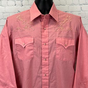 Può includere: Una camicia corallo con bottoni, a maniche lunghe e due tasche sul petto. La camicia presenta ricami floreali sulle spalle e sul colletto. La camicia ha chiusure a pressione perlata.