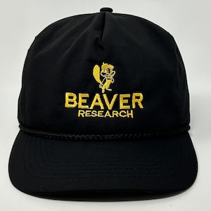 May include: Black baseball cap with a flat brim and a rope detail. The cap features the embroidered text "BEAVER RESEARCH" in gold, along with a cartoon beaver logo. The cap has a curved brim and a button on top.