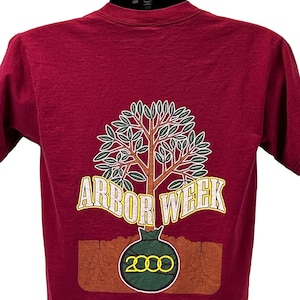 Puede incluir: Camiseta granate con un gráfico de árbol y el texto "ARBOR WEEK" en amarillo. El árbol tiene hojas verdes y ramas marrones. El año "2000" está impreso debajo en un círculo verde.