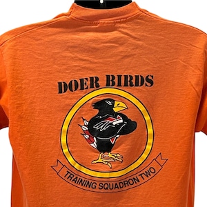 May include: An orange t-shirt displaying the text "DOER BIRDS" above a circular emblem. The emblem features a black bird with a yellow beak and red details, within a yellow border. The text "TRAINING SQUADRON TWO" is below the emblem.