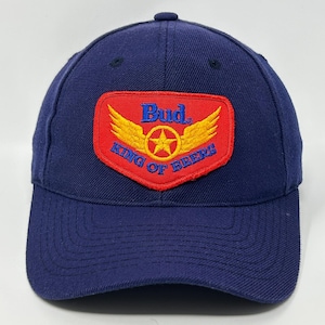 Puede incluir: Gorra de béisbol azul marino con visera curva. La gorra presenta un parche rojo con el texto "Bud. King of Beers" y un diseño de estrella y alas doradas. La gorra está hecha de tela texturizada.