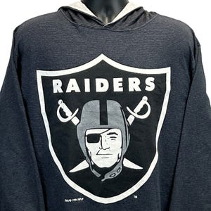 May include: Black and gray striped hooded sweatshirt featuring the Raiders logo. The logo is a shield shape with the word "RAIDERS" in white, a helmeted face, and crossed swords. The sweatshirt has a gray hood lining.