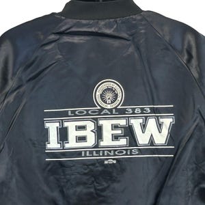 May include: A navy blue bomber jacket with a black collar. The back of the jacket features the text "LOCAL 383 IBEW ILLINOIS" in white block letters, with a circular emblem above. The jacket is made of a shiny material.