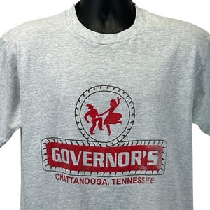 May include: A light gray t-shirt featuring a red graphic of two dancing figures inside a circular border. Below the graphic is the word "GOVERNOR'S" in red, with "CHATTANOOGA, TENNESSEE" underneath. The shirt has a crew neck.