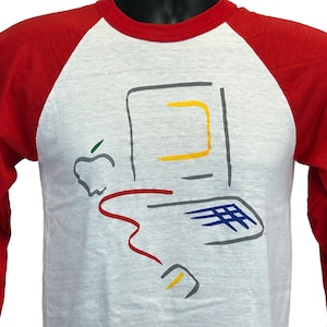 May include: A white and grey raglan-style shirt with red sleeves. The shirt features a colourful graphic of a vintage computer and the Apple logo. The computer graphic includes a yellow screen, a blue keyboard, and a red cable.