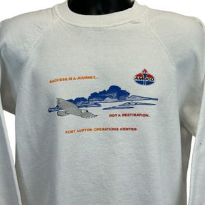Puede incluir: Sudadera blanca de cuello redondo con un gráfico de un pájaro volando, nubes azules y el logotipo de Amoco. El texto dice: "SUCCESS IS A JOURNEY... NOT A DESTINATION. FORT LUPTON OPERATIONS CENTER."