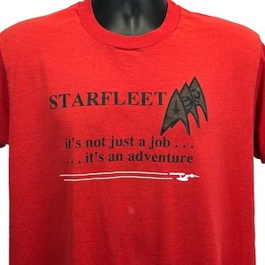 May include: Red Starfleet t-shirt with black text and graphics. The text reads "STARFLEET" above the phrase "it's not just a job... ...it's an adventure". The shirt features a Starfleet emblem.