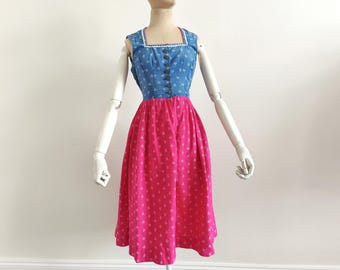 Puede incluir: Un vestido de dirndl rosa y azul con ribete blanco. El corpiño es de mezclilla azul con botones blancos y la falda es rosa con un estampado floral blanco.