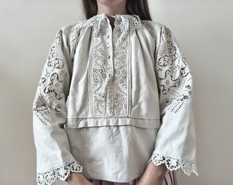 May include: A light beige long-sleeved blouse featuring intricate white lace panels on the chest and shoulders. The garment has a high collar, wide sleeves with lace-trimmed cuffs, and a central vertical lace insert extending from the neckline.