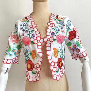 Könnte beinhalten: Eine weiße, kurze Jacke mit einem gewellten Saum und bunter Blumenstickerei. Die Stickerei zeigt rote, rosa, gelbe, blaue und grüne Blumen.