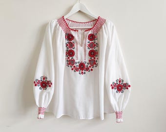 May include: White blouse with red floral embroidery. The long-sleeved top features detailed embroidery around the neckline, chest, and sleeves. The cuffs and neckline have a red zig-zag trim. The blouse is hanging on a white hanger.