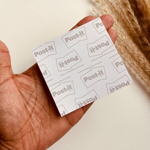 May include: A square Post-it note with a repeating pattern of the "Post-it" brand logo in gray on a white background. The words "Custom Printed Post-it Note" are also printed on the note. The note is held in a hand.
