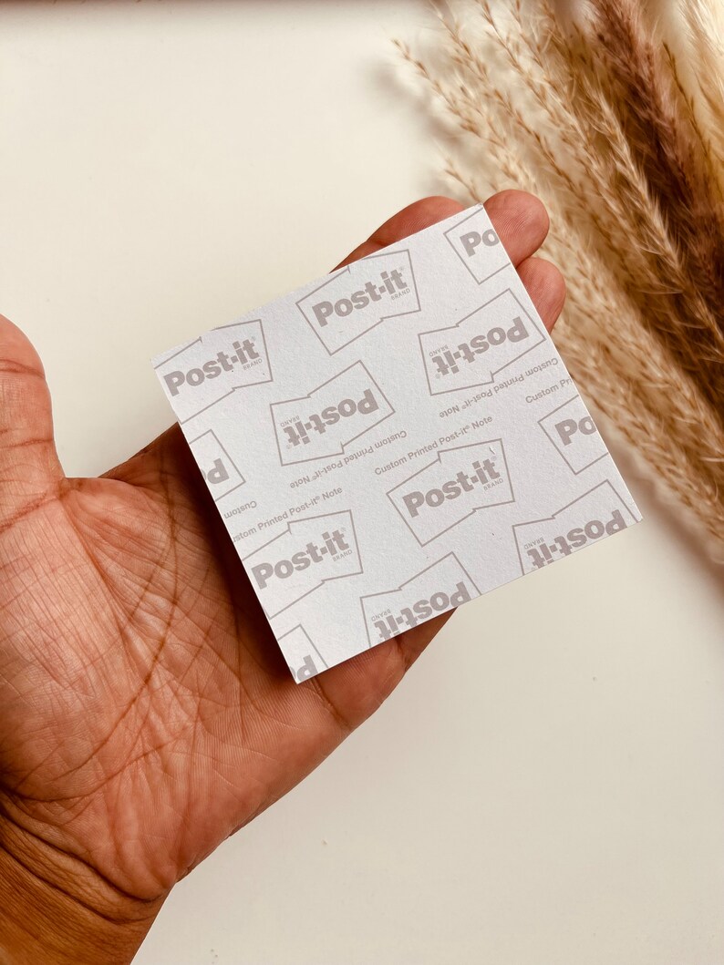 May include: A square Post-it note with a repeating pattern of the "Post-it" brand logo in gray on a white background. The words "Custom Printed Post-it Note" are also printed on the note. The note is held in a hand.