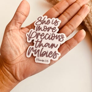 May include: A white sticker with the text "She is more precious than rubies" in brown cursive lettering. The text "Proverbs 3:15" is also included in smaller font below the main text.
