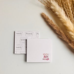 May include: A white Post-it note with the words "YOU ARE enough" in pink and red script. A partially visible Post-it pad backing is behind the note. Dried ornamental grass is in the background.