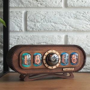 May include: A wooden clock with a retro design features four Nixie tubes displaying the time. The clock has a round knob and the text "Greenwich" on the bottom.