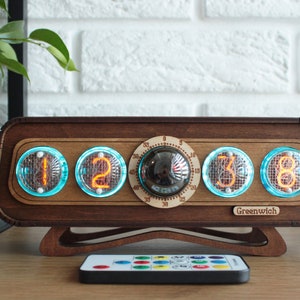 May include: A wooden clock with four orange digital displays showing the time 12:38. The clock has a round knob and the text "Greenwich" on the bottom. A remote control with colored buttons is in the foreground.