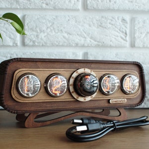 May include: A wooden clock with a retro design features four red glowing Nixie tubes displaying the time. The clock has a round knob for setting the time and the text "Greenwich" on the front. The clock is sitting on a wooden surface with a black power cord.