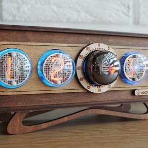 May include: A wooden clock with four Nixie tube displays showing the time 12:16. The clock has a knob for setting the time and a label that says "Greenwich".