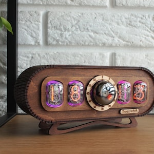 May include: A wooden clock with a vintage style featuring four Nixie tubes displaying the time. The clock has a round knob for setting the time and the text "Greenwich" printed on the front.