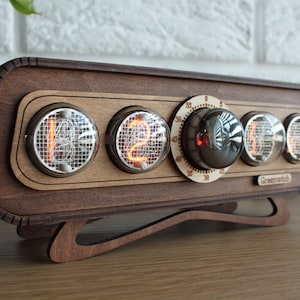 May include: A wooden clock with a retro design features four Nixie tubes displaying the time "12" and a round knob with numbers from 0 to 60. The clock is labeled "Greenwich" and sits on a wooden base.