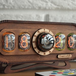 May include: A wooden clock with a retro design features four orange and green Nixie tubes displaying the time "18:20". The clock has a round knob with a silver finish and a white dial with black markings. The clock is labeled "Greenwich" and is sitting on a wooden surface with a remote control nearby.