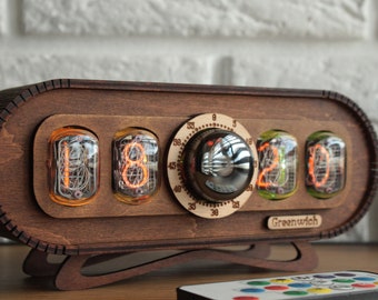 May include: A wooden clock with a retro design features four orange and green Nixie tubes displaying the time "18:20". The clock has a round knob with a silver finish and the text "Greenwich" on the bottom. A remote control with colorful buttons is in the foreground.