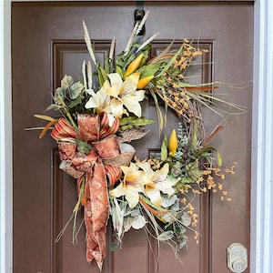 May include: A fall-themed wreath with a brown base, featuring a large bow with orange, red, and gold accents. The wreath is decorated with yellow and white lilies, green leaves, and brown and yellow grasses.