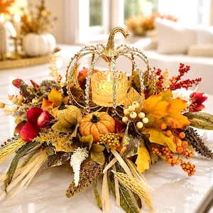 May include: A decorative centerpiece featuring a gold-toned wire pumpkin frame encrusted with clear crystals, surrounding a glass votive candle holder. The arrangement includes artificial orange pumpkins, autumn leaves, red berries, and dried wheat stalks.