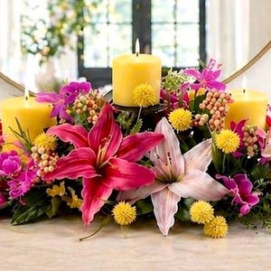 May include: A floral centerpiece with three lit yellow candles. The arrangement includes vibrant pink and purple lilies, yellow pom-poms, and greenery. The candles are set on a table with a mirror in the background.