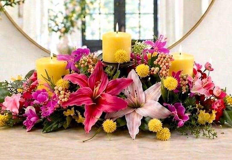 May include: A floral centerpiece with three lit yellow candles. The arrangement includes vibrant pink and purple lilies, yellow pom-poms, and greenery. The candles are set on a table with a mirror in the background.