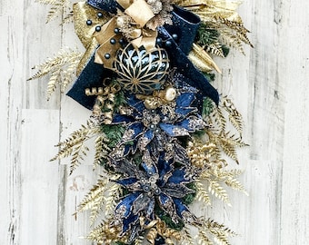 Puede incluir: Una guirnalda navideña azul y dorada con un gran adorno azul y dorado, una poinsettia azul y dorada y un adorno en forma de lágrima azul y dorado.
