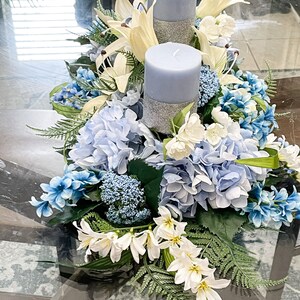 Puede incluir: Un centro de mesa con dos velas azules con detalles de purpurina plateada rodeadas de flores blancas y azules. Las flores incluyen hortensias, lirios y helechos.