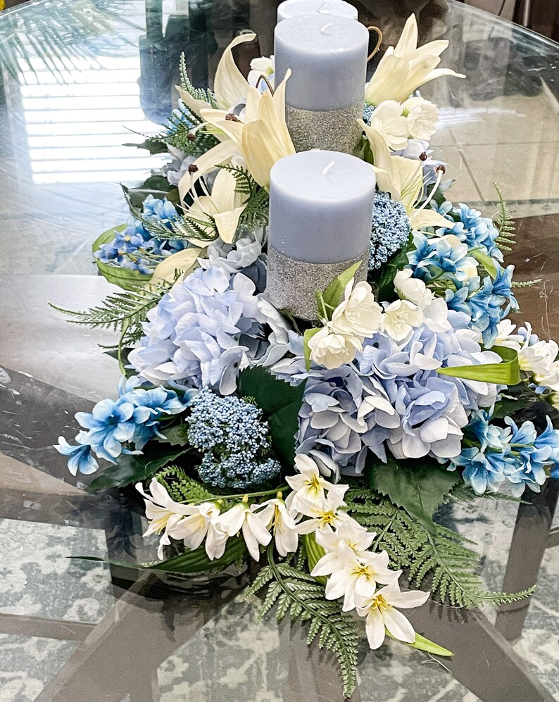Puede incluir: Un centro de mesa con dos velas azules con detalles de purpurina plateada rodeadas de flores blancas y azules. Las flores incluyen hortensias, lirios y helechos.
